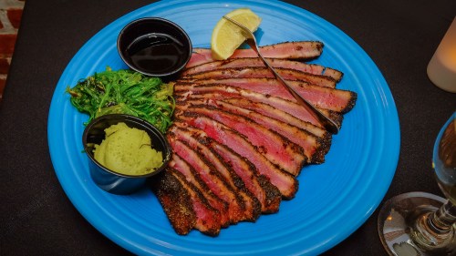 Sliced grilled meat on a blue plate, accompanied by wasabi, seaweed salad, and dipping sauce, garnished with lemon.