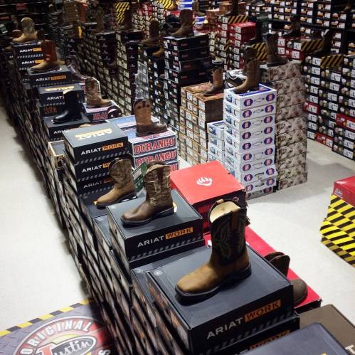 Aisle filled with various brands of cowboy boots displayed on boxes in a retail store.