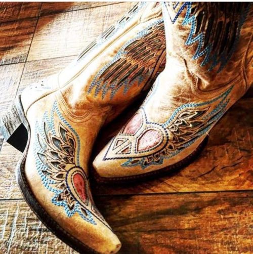 A close-up of stylish cowboy boots with intricate wing designs in blue and pink on a wooden floor.