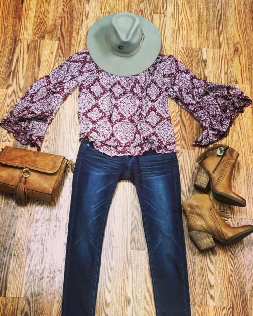 Flat lay of a patterned blouse, jeans, cowboy hat, brown handbag, and heeled boots on a wooden surface.