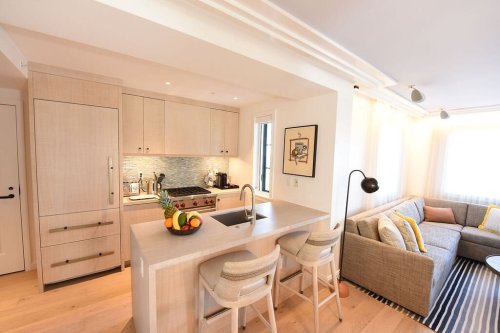 Modern kitchen and living area with light wood finishes, a fruit bowl, and a cozy sofa with colorful cushions.