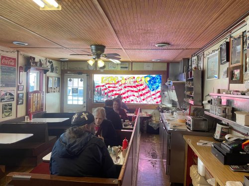 A cozy diner interior with patrons at tables, vintage decor, and a large American flag displayed in the window.