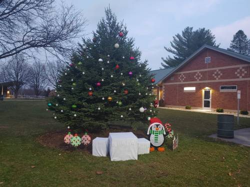 A decorated Christmas tree with colorful ornaments stands in a park, next to a snowman decoration and a building.