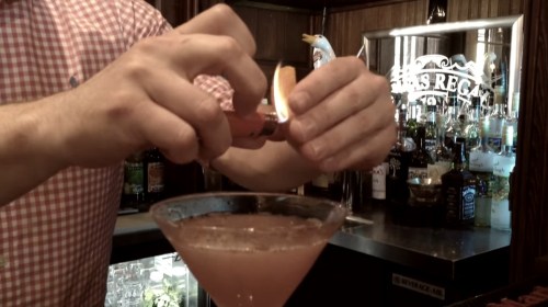 A bartender lights a citrus peel over a cocktail in a glass, with a bar full of bottles in the background.