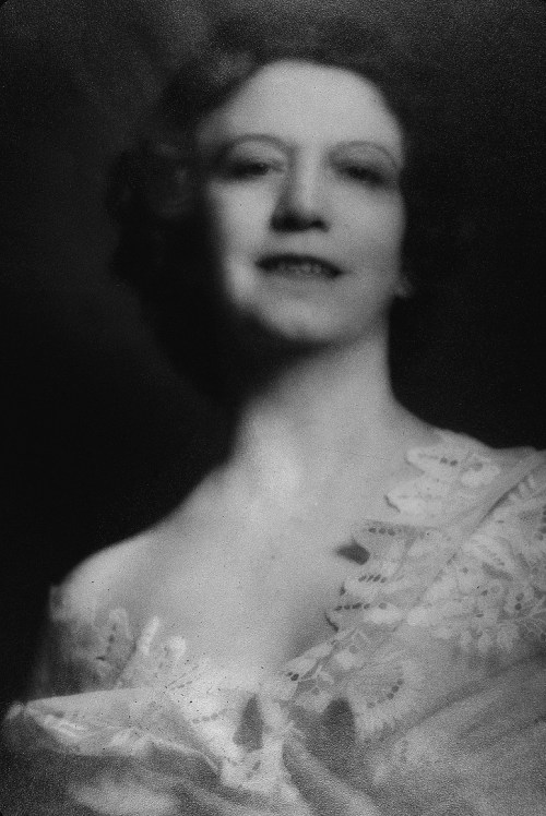A vintage black-and-white portrait of a smiling woman with wavy hair, wearing a lace dress.
