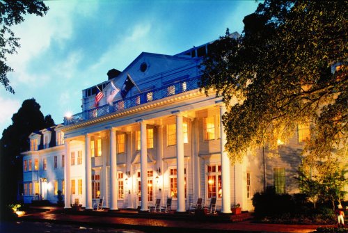 A grand white mansion illuminated at dusk, featuring columns, flags, and a welcoming porch with rocking chairs.