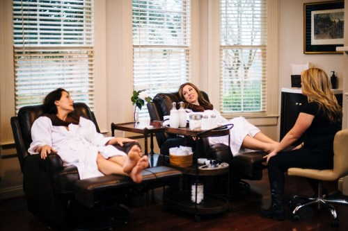 Two women in robes relax in massage chairs while a third woman attends to them in a cozy spa setting.