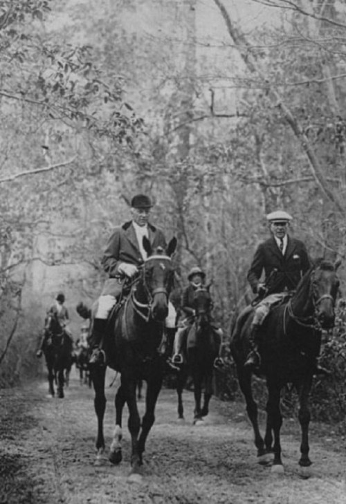 Black and white photo of a group of people on horseback riding through a wooded path.