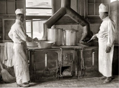 Two chefs in white uniforms cooking in a vintage kitchen with large pots and an old-fashioned stove.
