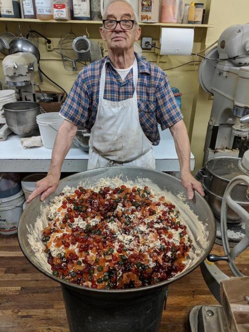 A man in an apron stands behind a large bowl filled with colorful fruit and dough mixture in a bakery.