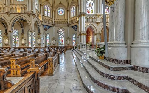 Interior of a grand church featuring stained glass windows, wooden pews, and marble steps leading to an altar.