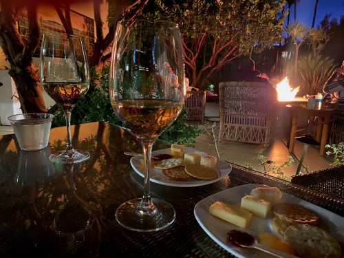 Two glasses of wine beside plates of cheese and crackers, with a cozy outdoor fire and greenery in the background.
