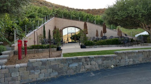 A modern building with outdoor seating, surrounded by greenery and a stone wall, set against a hillside.