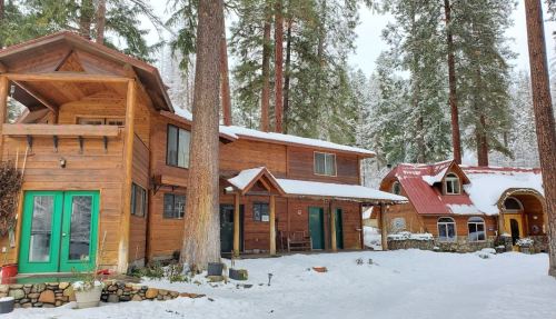 Two wooden cabins surrounded by snow and tall trees, with a cozy, rustic appearance in a winter landscape.