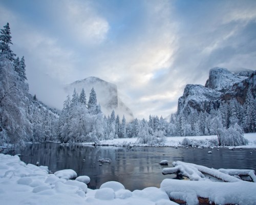 A serene winter landscape featuring snow-covered trees, a calm river, and mountains under a cloudy sky.