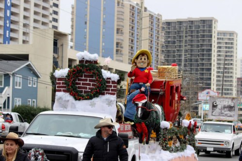 A festive parade float featuring a cowboy figure on a horse, decorated with holiday elements and surrounded by buildings.