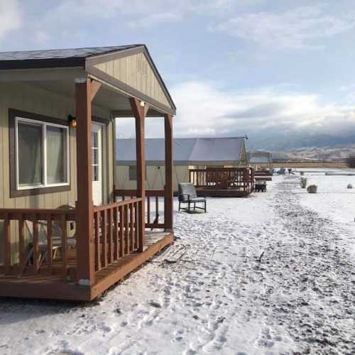 A small cabin with a porch in a snowy landscape, surrounded by other buildings and mountains in the background.