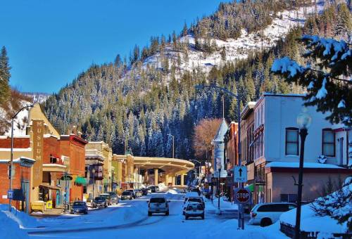 Snow-covered street in a small town, with colorful buildings and a backdrop of pine-covered mountains.
