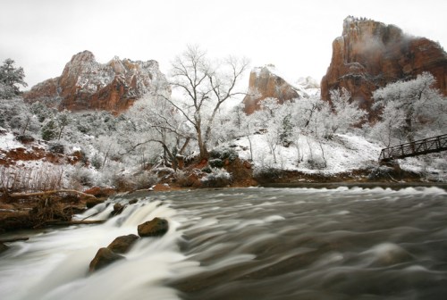 A snowy landscape with a flowing river, rocky cliffs, and bare trees in a serene, winter setting.