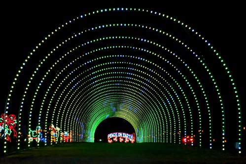 A colorful light tunnel with green and white lights, featuring festive decorations and a "Peace on Earth" sign.