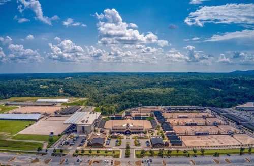 Aerial view of a large facility with multiple buildings and outdoor arenas, surrounded by green hills and a blue sky.
