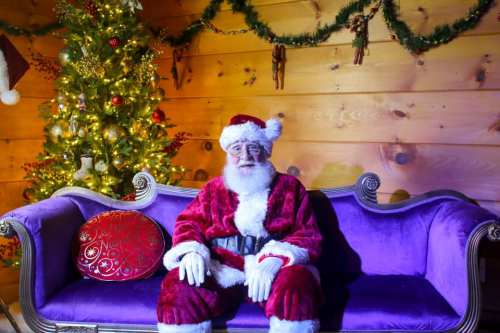 Santa Claus in a red suit sits on a purple couch, with a decorated Christmas tree in the background.