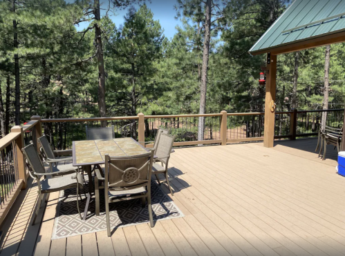 A wooden deck surrounded by trees, featuring a table with chairs and a rug, under a clear blue sky.