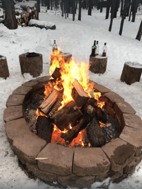 A cozy campfire surrounded by stone seating, with snow-covered trees in the background. Bottles are placed nearby.