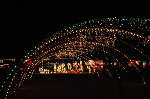 A festive display of colorful lights arches over a scene with illuminated figures, creating a cheerful holiday atmosphere.