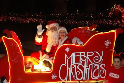Santa Claus and a woman in a Santa hat wave from a festive sleigh decorated with "Merry Christmas" lights.