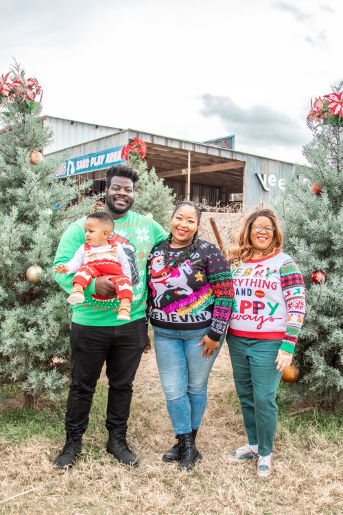 A family poses together in festive sweaters among Christmas trees, smiling and holding a baby.
