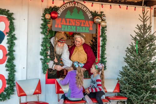 Two adults in festive attire interact with children at a holiday-themed booth, surrounded by Christmas decorations.