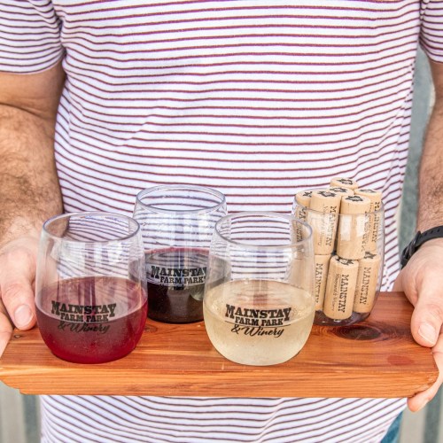A person holds a wooden tray with three wine glasses and corks from Mainstay Farm Park & Winery.