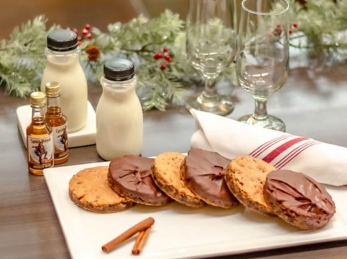 A plate of chocolate-dipped cookies with milk, mini liquor bottles, and festive decorations on a table.