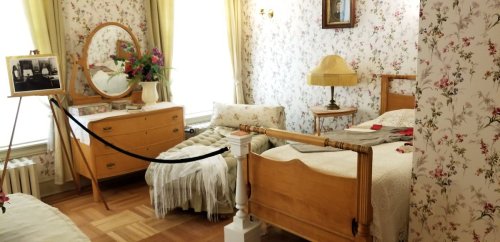 A vintage bedroom with floral wallpaper, a wooden bed, dresser, mirror, and a cozy armchair.