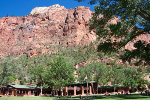 A lodge nestled at the base of towering red rock cliffs, surrounded by green trees under a clear blue sky.