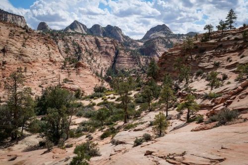 A scenic view of rugged red rock formations and green trees under a partly cloudy sky in a mountainous landscape.