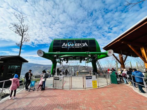 A ski lift station with a green structure, people waiting in line, and a scenic view under a partly cloudy sky.