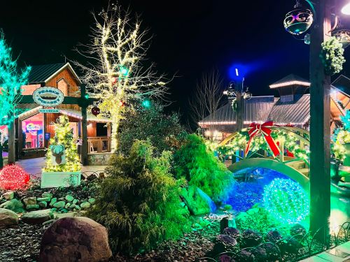 Colorful holiday lights illuminate a festive scene with a bridge, trees, and decorated buildings at night.