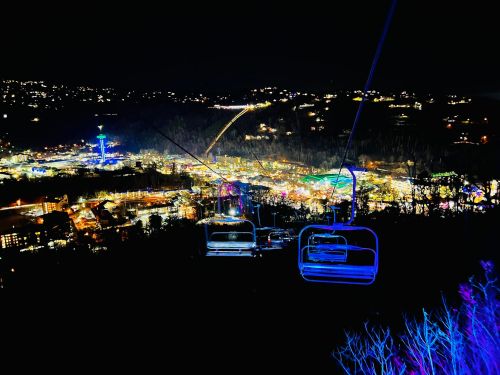 A night view from a ski lift overlooking a brightly lit town with colorful lights and hills in the background.