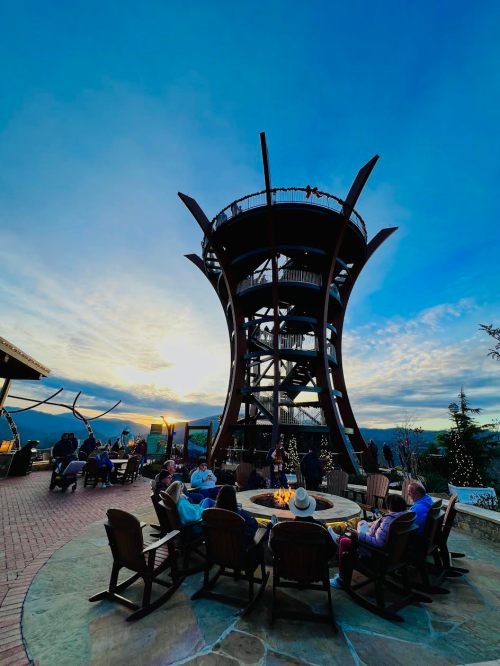 A scenic outdoor area with a tall structure, surrounded by people sitting around a fire pit at sunset.