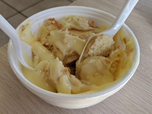 A bowl of dessert with cake pieces and creamy custard, two plastic spoons resting inside.