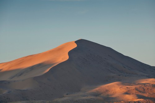 A sunlit sand dune with a smooth, curved peak against a clear blue sky.