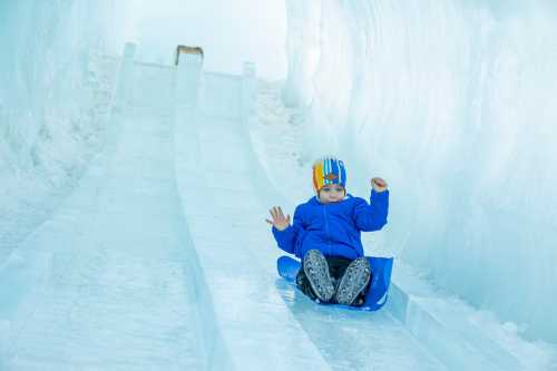 A child in a blue jacket sleds down a snowy ice slide, smiling with hands raised in excitement.