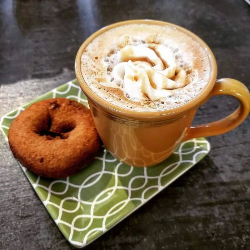 A cup of frothy coffee topped with whipped cream, alongside a chocolate donut on a green patterned plate.