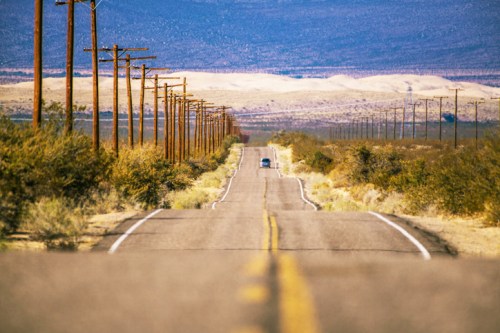 A long, straight road stretches into the distance, flanked by power lines and desert landscape under a clear sky.