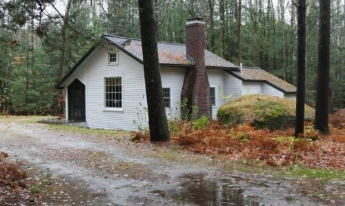 A quaint white house surrounded by trees and autumn foliage, with a gravel driveway leading up to it.