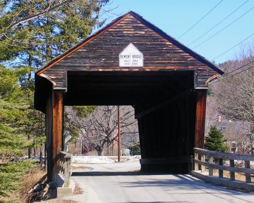 A wooden covered bridge with a sign reading "Bennett Bridge" spans a road, surrounded by trees and a clear blue sky.