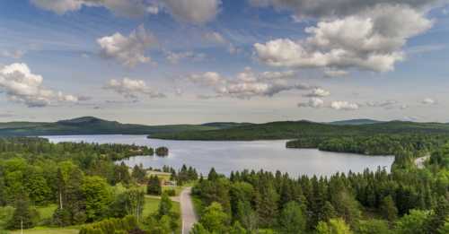 A serene landscape featuring a calm lake surrounded by lush green forests and rolling hills under a cloudy sky.