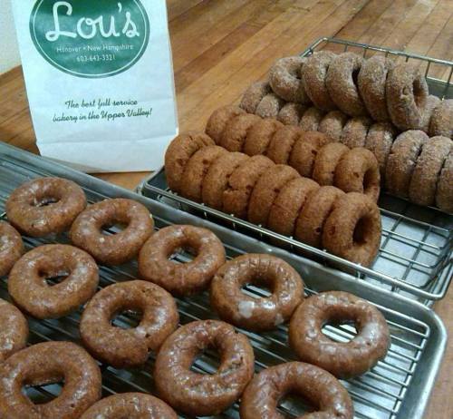A display of freshly made donuts on trays, with a bag from Lou's in the background.